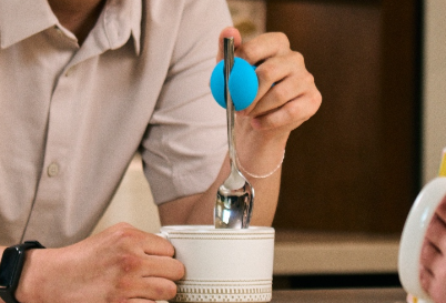a man is smiling as he holds a spoon in a medium grip and dips the spoon into a mug. He leans on a counter across from a woman holding a mug