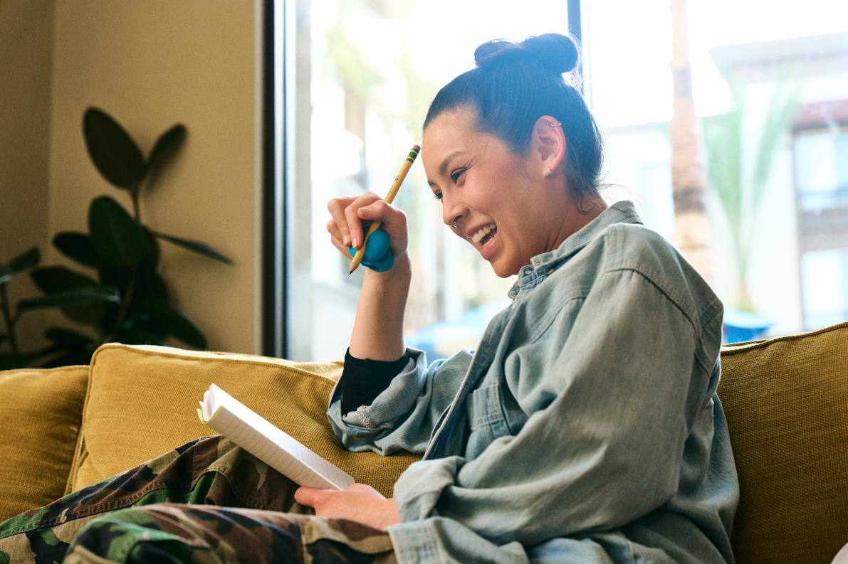 a girl sits on a yellow couch holding a pencil in a medium blue grip in one hand, and a notebook in the other, smiling